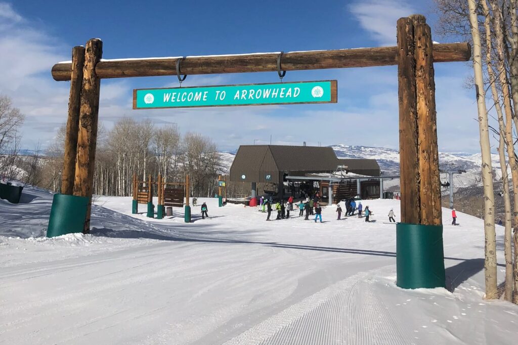 Timber gateway with a Welcome to Arrowhead sign above a groomed run, with the chairlift base and ski school groups behind