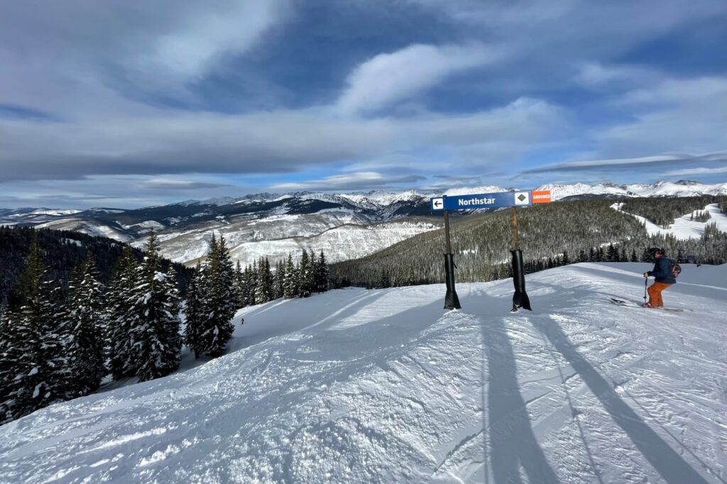 Skier beside a groomed blue run sign high on Vail Mountain, with the snow-covered Gore Range on the horizon